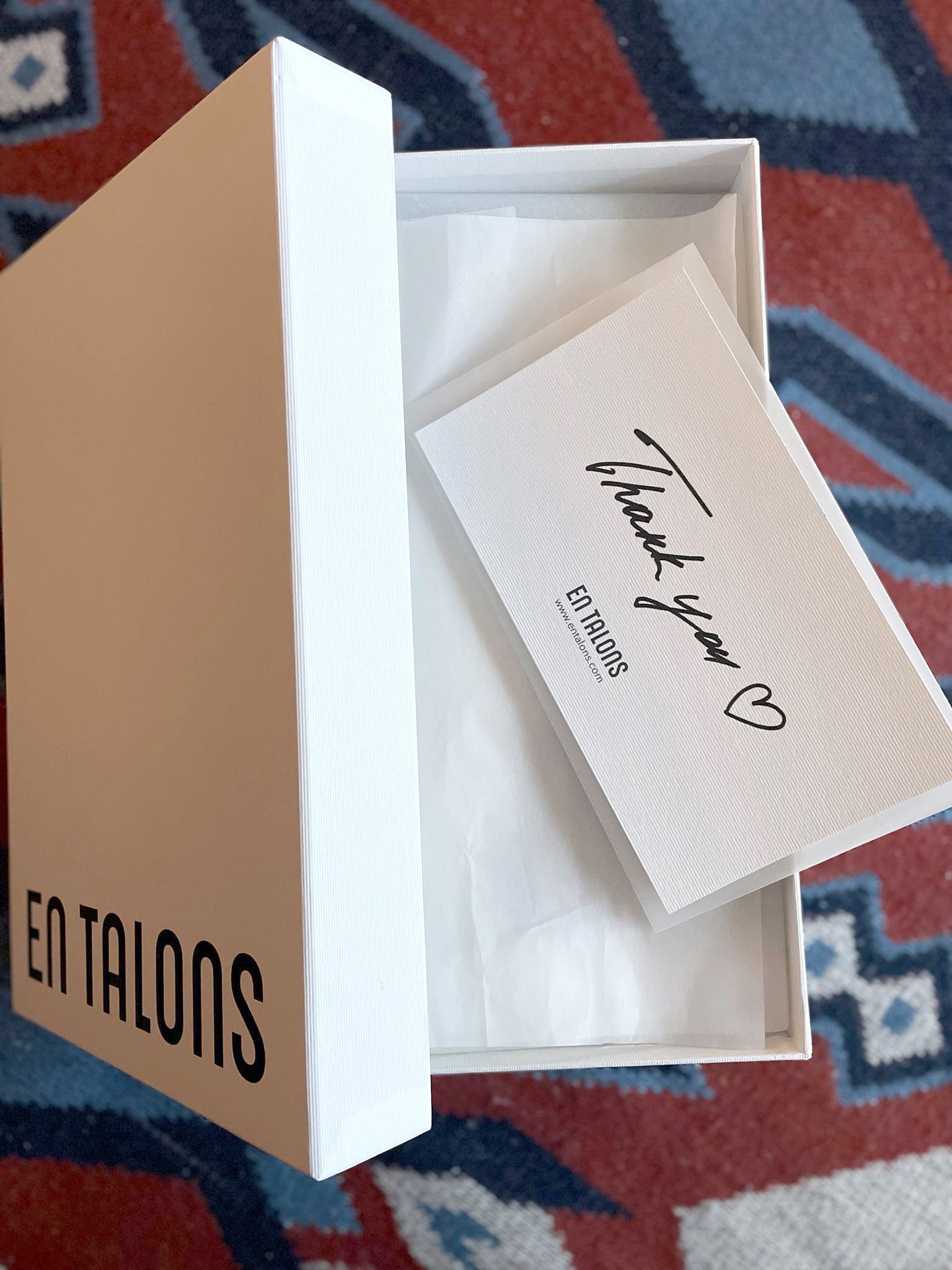 A white gift box with the lid off, showing a white tissue insert and a folded thank-you card with 'Thank you' in script and 'EN TALONS' printed on it. The box is placed on a red and blue patterned rug.