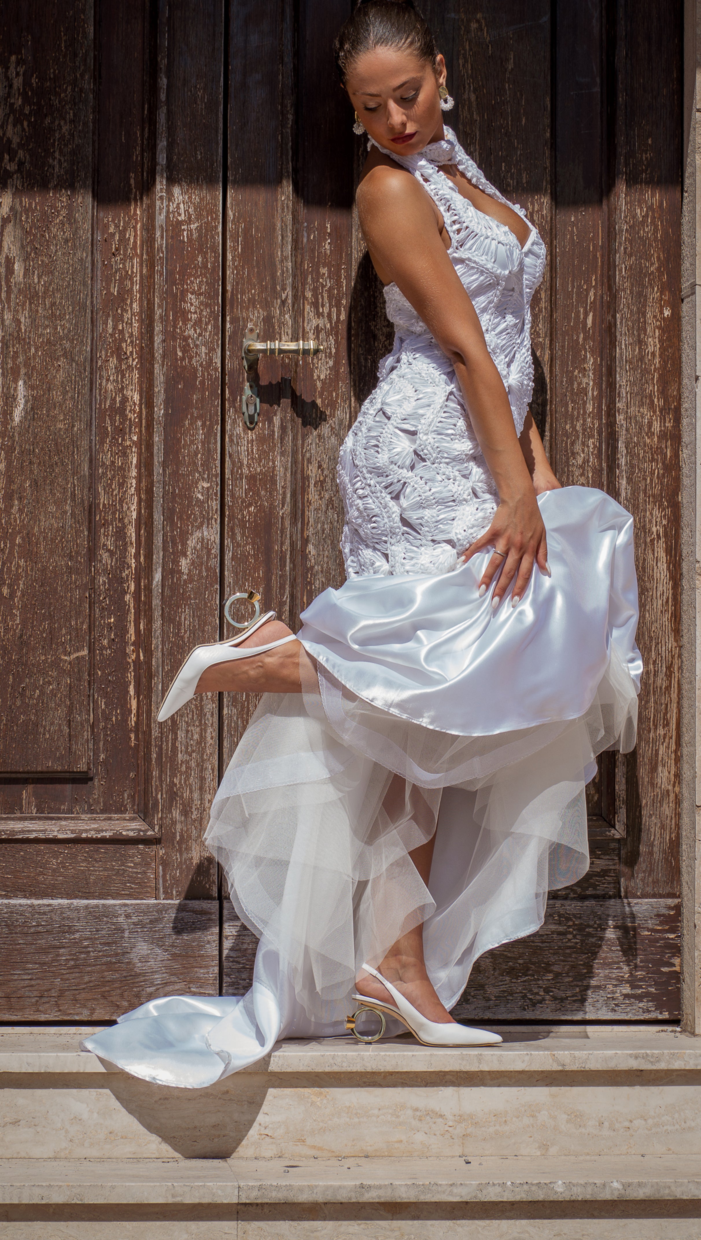 A woman in a white wedding gown poses against a rustic dark wooden door, holding up the skirt to show La Bague slingback heels.
