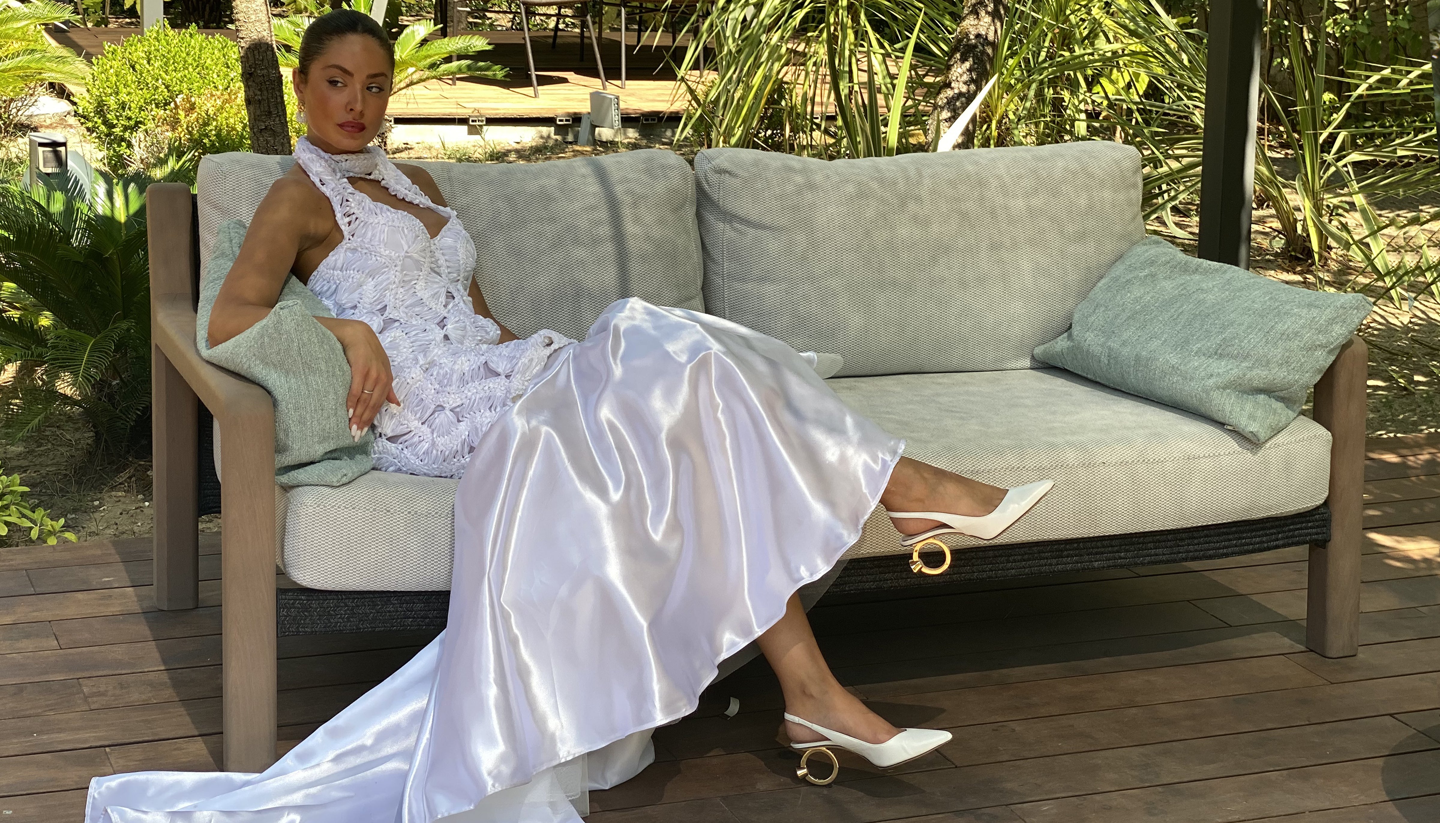 A woman reclining on a cushioned outdoor sofa wearing a wedding dress with a flowing satin train and white pointed slingback with gold La Bague heels , set on a wooden floor.