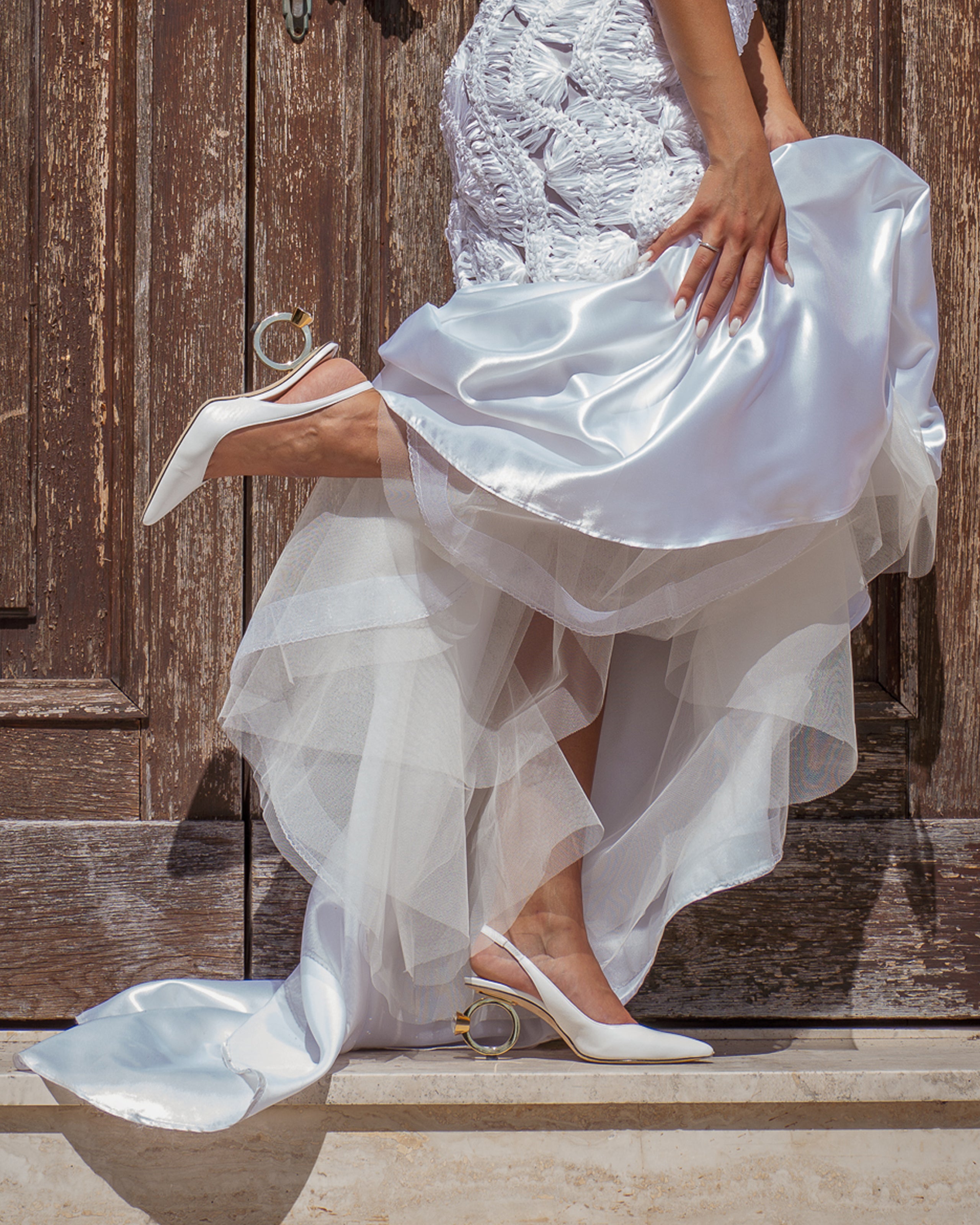 A woman seated against a rustic wooden door wearing a wedding dress with a flowing satin and tulle skirt and white slingback with La Bague heels.