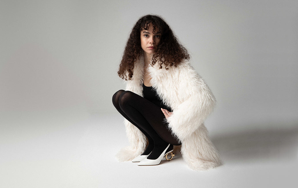 Model wearing white slingbacks with jeweled heels, spotlight emphasizing the elegant design.
